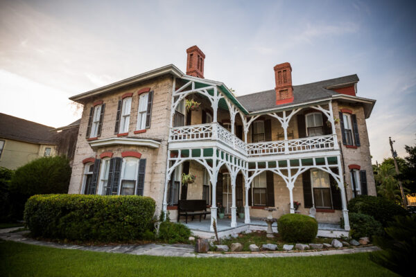 A two-story historic house with ornate wooden trim, red brick chimneys, and a wraparound porch sits among manicured bushes and a green lawn under a partly cloudy sky.