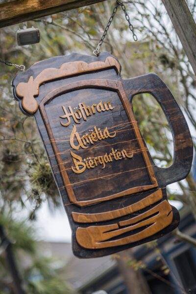 Wooden sign shaped like a beer mug with the words Hofbrau Amelia Biergarten carved in gold script, hanging outdoors by metal chains, with trees in the background.