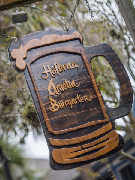 A wooden sign shaped like a beer stein hangs outdoors, featuring the words Hofbräu Amelia Biergarten in gold lettering, with trees and foliage blurred in the background.