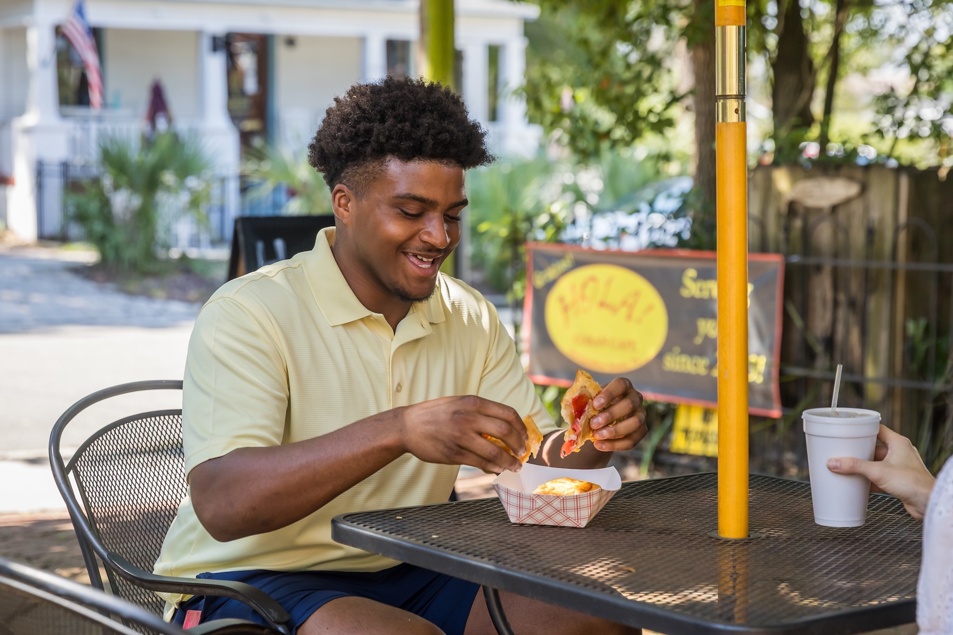 A man sits at an outdoor table, smiling as he eats a sandwich and fries from a food basket. Another person’s hand holds a cup across the table. Trees and a building are visible in the background.
