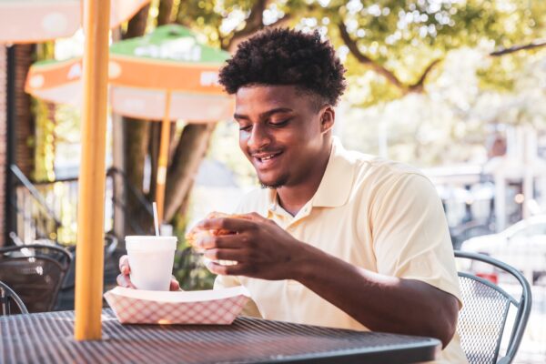A young man sitting at an outdoor café table, smiling while holding a sandwich and a cup with a straw. Sunlight filters through the trees and colorful umbrellas are in the background.