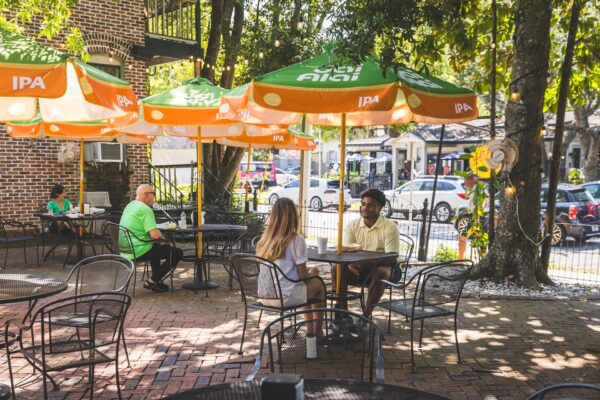 Two people sit at an outdoor café table under large green and orange umbrellas. Other patrons are nearby, and parked cars can be seen on the street in the background. The scene is shaded by trees.
