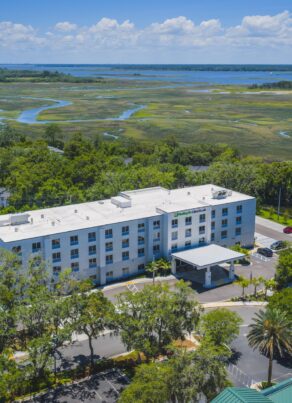 Aerial view of a modern white hotel surrounded by trees, with a marsh and waterway in the background under a partly cloudy sky.