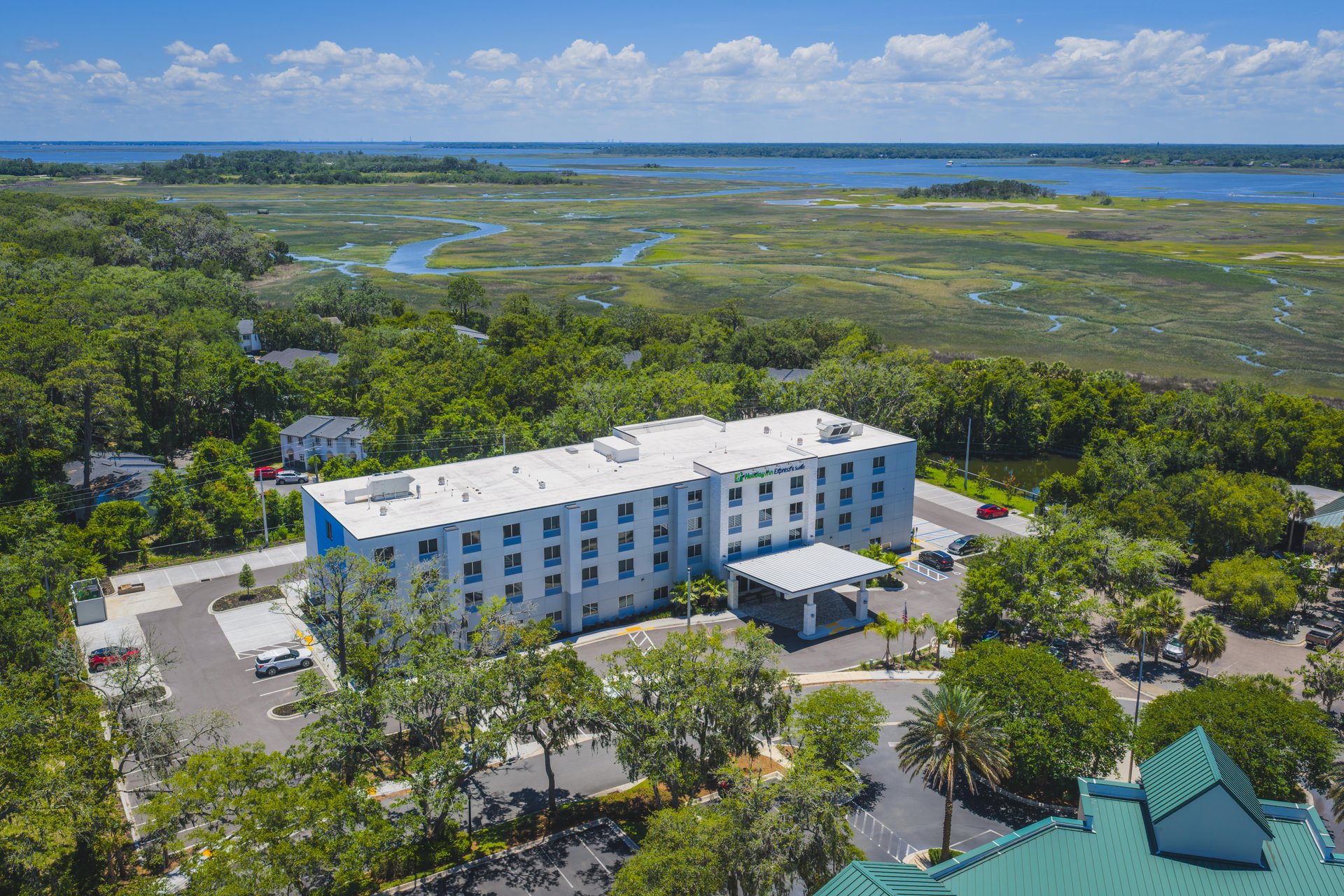 Aerial view of a modern white hotel surrounded by trees, with a marsh and waterway in the background under a partly cloudy sky.
