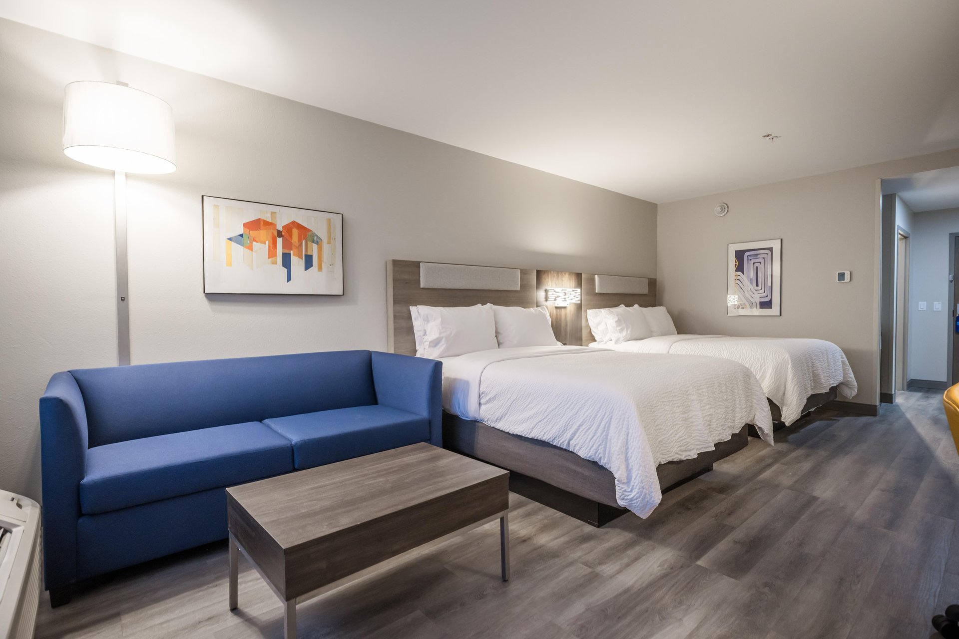 Hotel room with two beds, a blue sofa, wooden coffee table, wall art, standing lamp, and gray wood flooring under soft lighting. A hallway leads to another area.