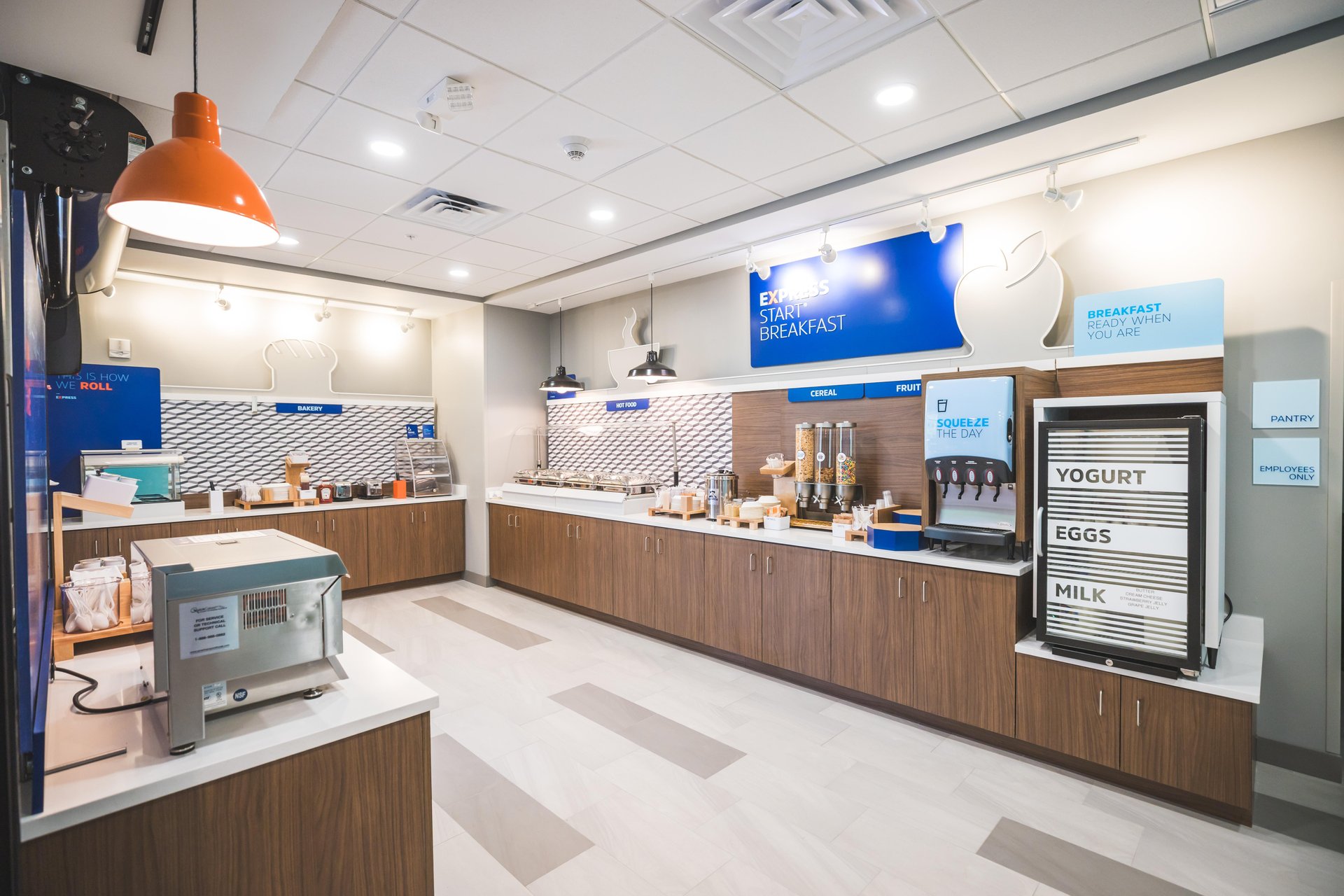 Modern hotel breakfast area with food stations, cereal dispensers, a drink machine, microwave, and a sign displaying options like yogurt, eggs, and milk. Bright lighting and clean, organized counters.