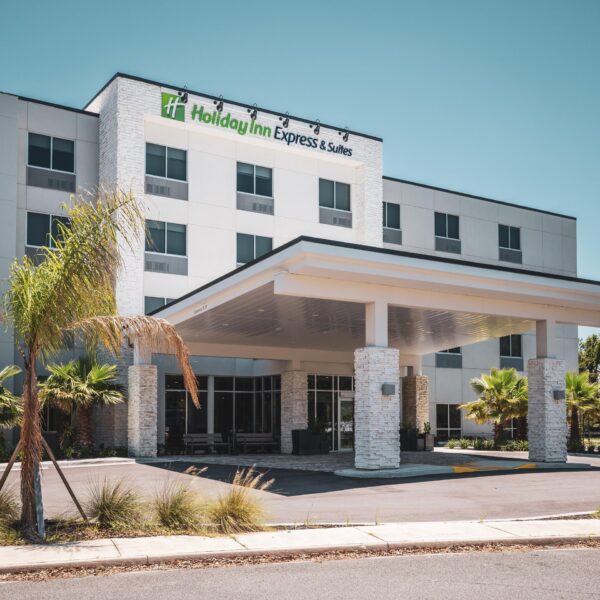 A modern Holiday Inn Express & Suites hotel with white stone exterior, palm trees, large front entrance canopy, and clear blue sky.