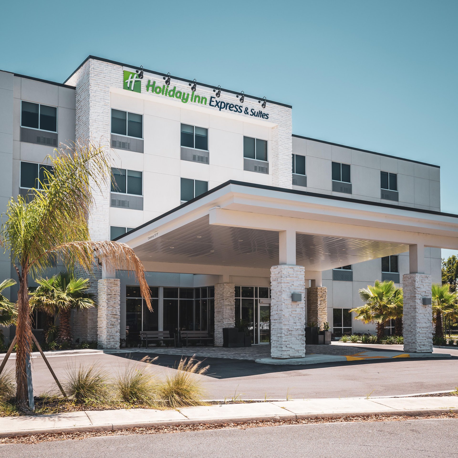 A modern Holiday Inn Express & Suites hotel with white stone exterior, palm trees, large front entrance canopy, and clear blue sky.