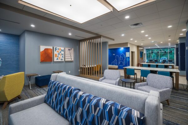 Modern lounge area with gray couches, patterned pillows, armchairs, tables, wall art, wood slats, and a long communal table. Large windows bring in natural light. Blue accents and cityscape mural on the walls.