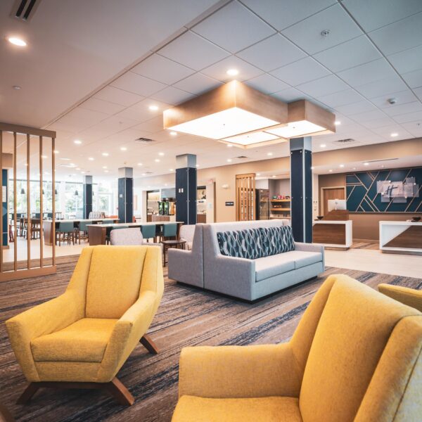 A modern hotel lobby with yellow armchairs, a gray sofa, stylish lighting, wood accents, and a reception desk in the background. Large windows provide natural light, and the space looks clean and inviting.