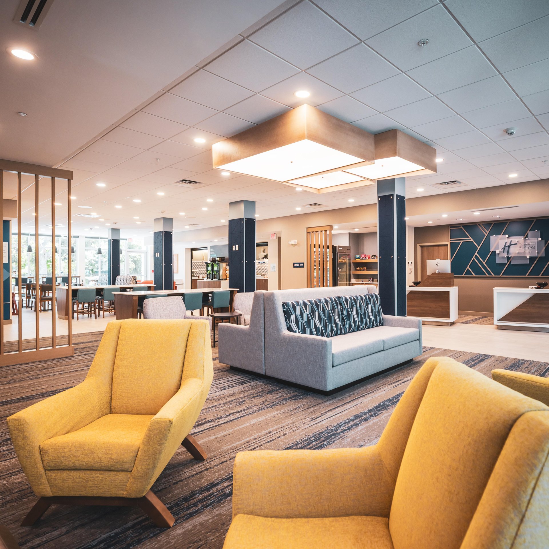 A modern hotel lobby with yellow armchairs, a gray sofa, stylish lighting, wood accents, and a reception desk in the background. Large windows provide natural light, and the space looks clean and inviting.
