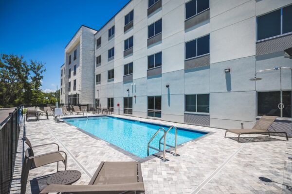 Outdoor hotel pool with clear blue water, surrounded by lounge chairs and tables, next to a modern four-story building with large windows on a sunny day.