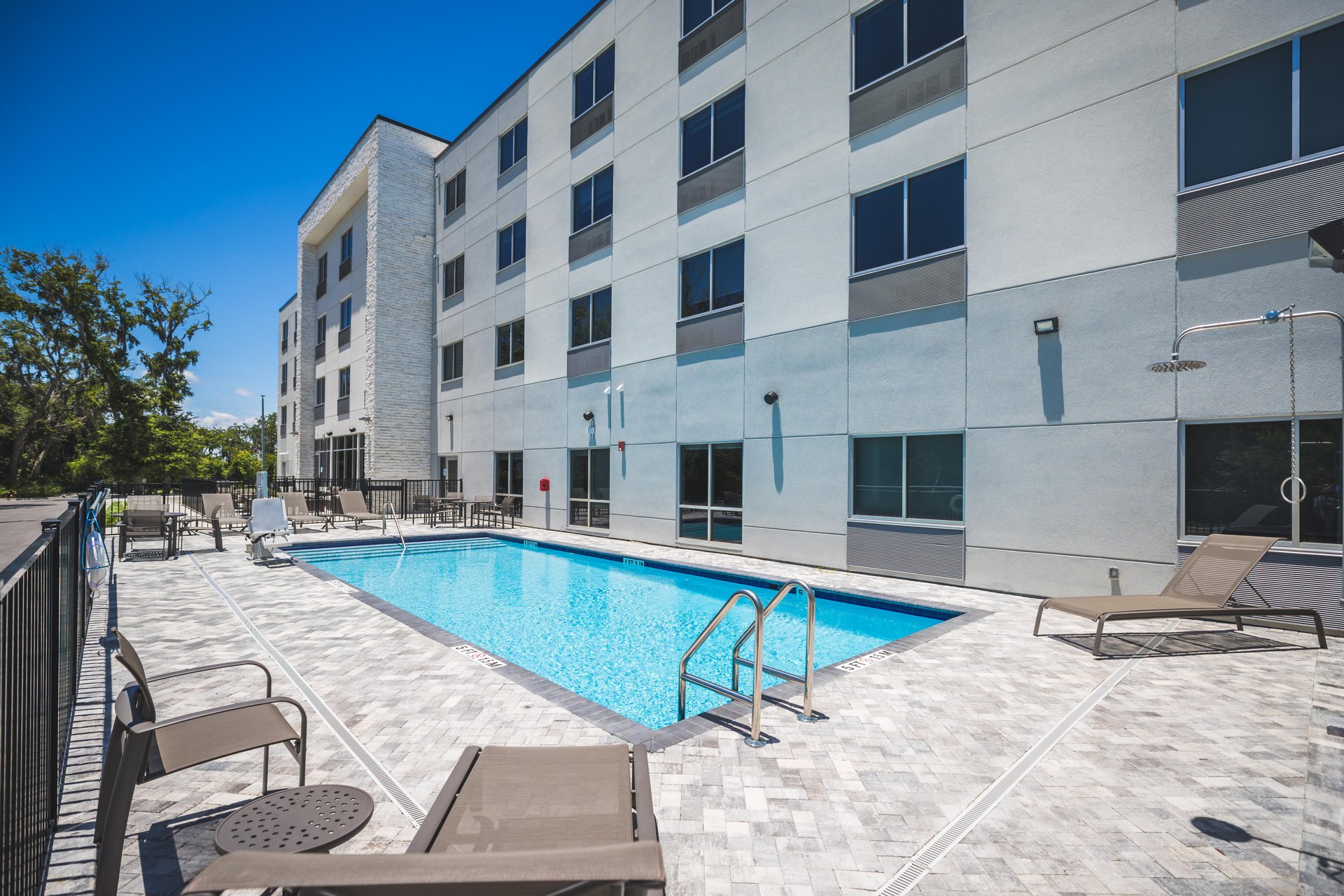 Outdoor hotel pool with clear blue water, surrounded by lounge chairs and tables, next to a modern four-story building with large windows on a sunny day.