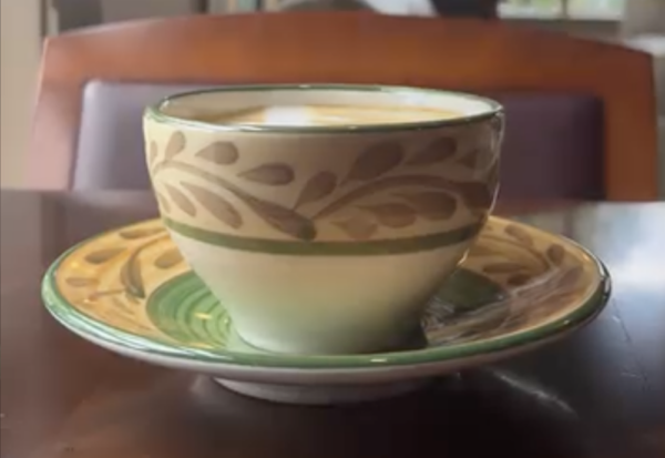 A decorative ceramic cup filled with coffee sits on a matching saucer with leaf patterns, placed on a dark wooden table in a softly lit café.