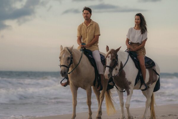 A man and a woman ride horses side by side along a sandy beach at sunset, with ocean waves in the background. The man wears a tan shirt and white pants; the woman wears a white shirt and tan shorts.