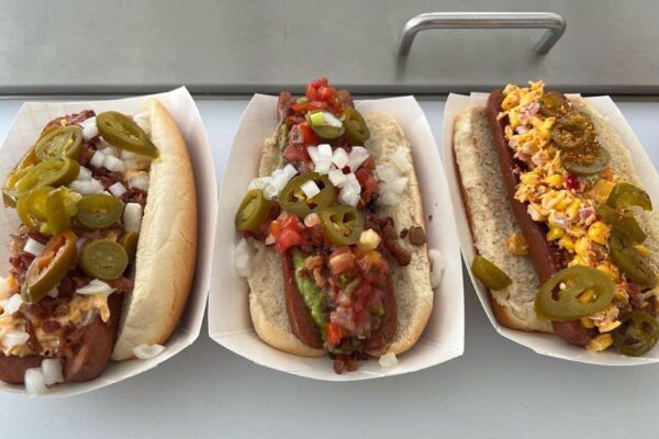 Three hot dogs in paper trays topped with various toppings: the left one with jalapeños, bacon, onions, and cheese; the middle with pico de gallo, guacamole, onions, and jalapeños; the right with cheese, bacon, and jalapeños.