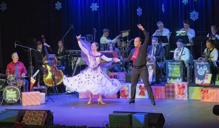 A woman in a flowing white dress and a man in a black outfit with a red scarf dance on stage, surrounded by Christmas decorations and gifts. A live band plays in the background under blue stage lights and hanging snowflakes.