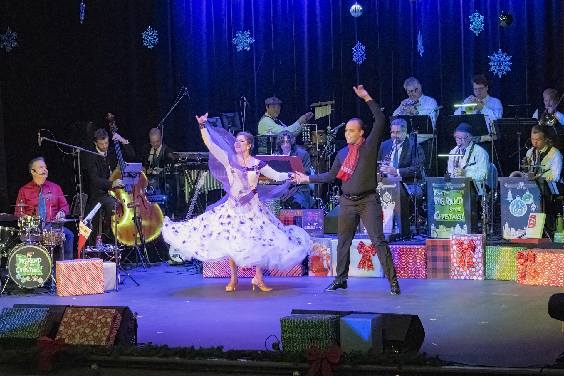 A pair dancing on a stage in front of a live big band, with Christmas-themed gift boxes arranged across the floor.