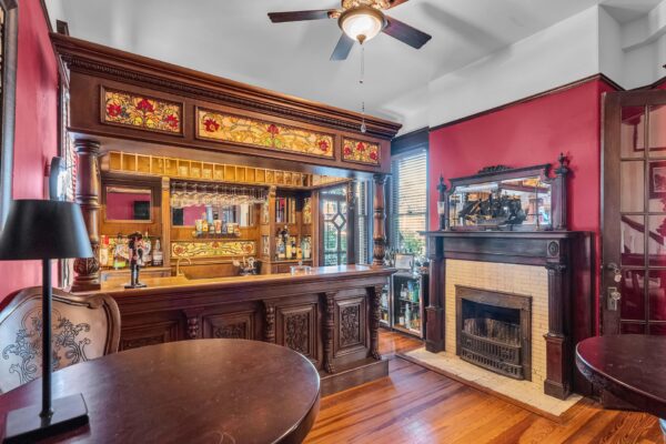 Victorian-style home bar with ornate woodwork, stained glass panels, shelves of bottles, a dark wooden bar counter, a ceiling fan, and a fireplace with a mirror above, all in a richly decorated room with red walls.
