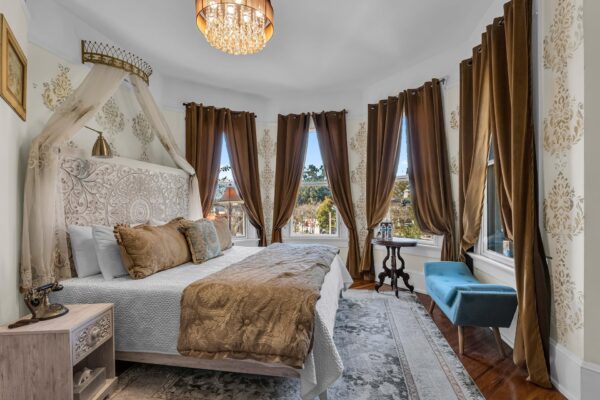 Elegant bedroom with a carved headboard, gold and white bedding, canopy, and brown curtains on tall windows. A chandelier hangs above, with a blue upholstered bench and vintage side tables completing the decor.