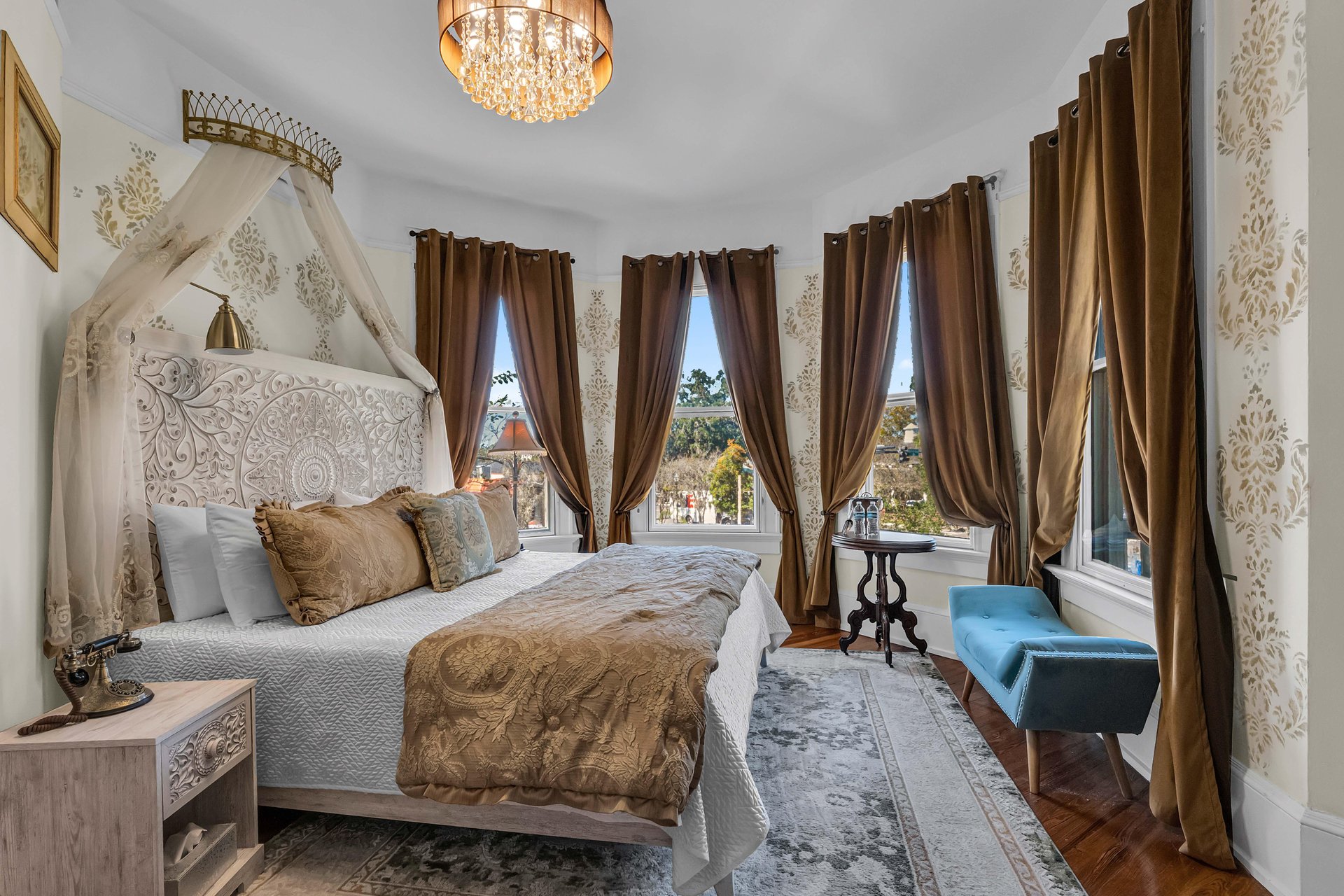 Elegant bedroom with a carved headboard, gold and white bedding, canopy, and brown curtains on tall windows. A chandelier hangs above, with a blue upholstered bench and vintage side tables completing the decor.