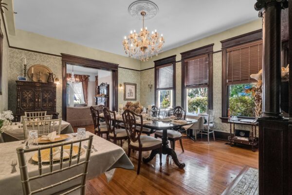 Elegant dining room with wooden floors, tables set for a meal, classic wooden chairs, large windows with brown blinds, a crystal chandelier, and ornate dark wood trim and furniture. Light streams in from outside.
