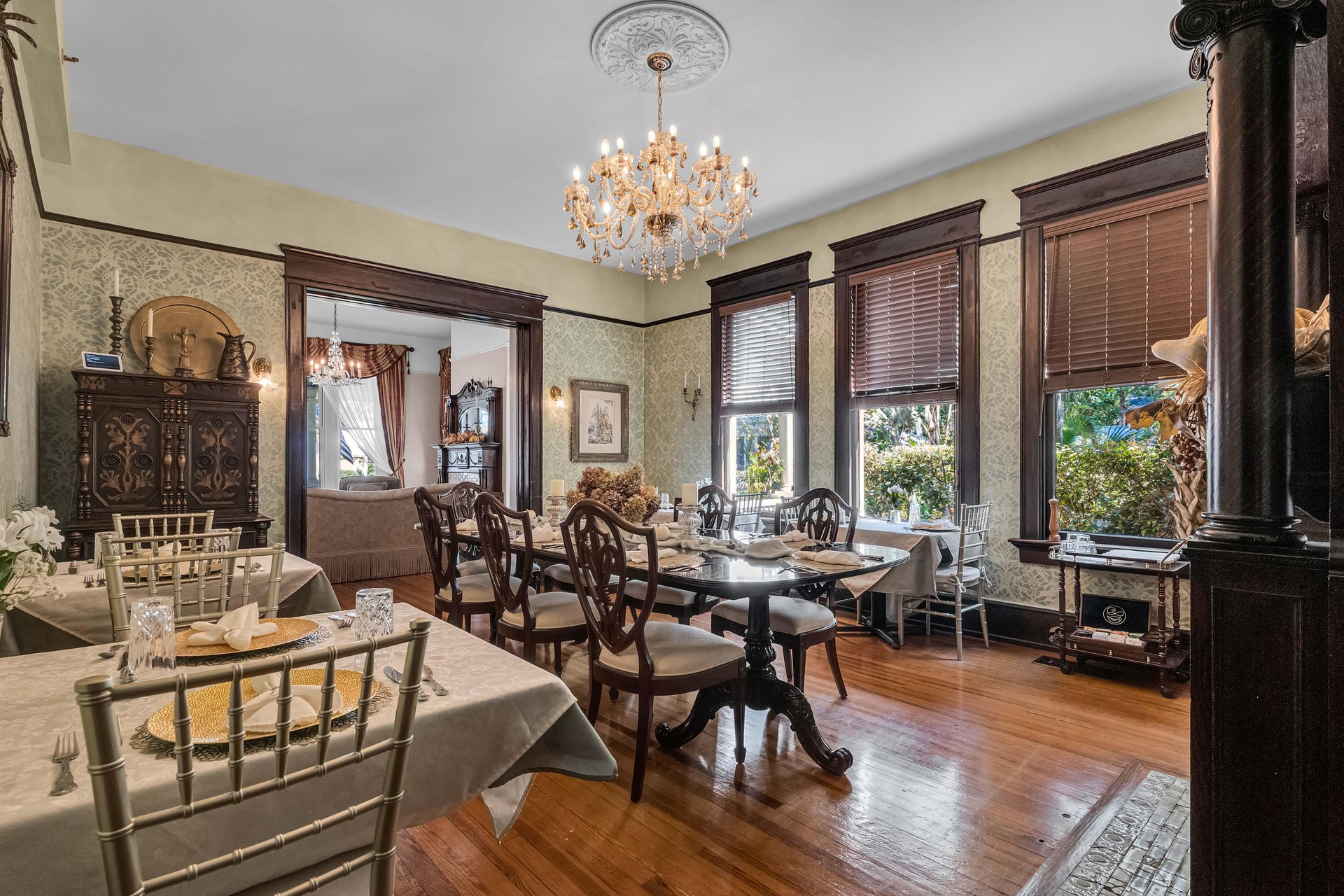 Elegant dining room with wooden floors, tables set for a meal, classic wooden chairs, large windows with brown blinds, a crystal chandelier, and ornate dark wood trim and furniture. Light streams in from outside.