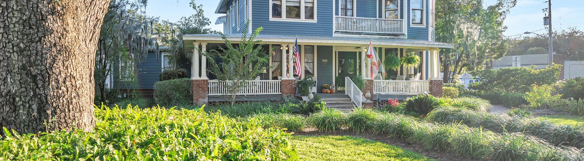 A large blue Victorian-style house with a wraparound porch sits behind lush green landscaping and a curved walkway. Mature trees with hanging moss frame the scene on a sunny day.
