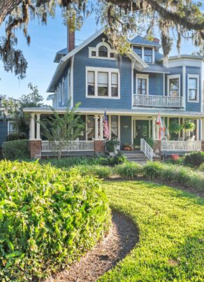 A large blue Victorian-style house with a wraparound porch sits behind lush green landscaping and a curved walkway. Mature trees with hanging moss frame the scene on a sunny day.