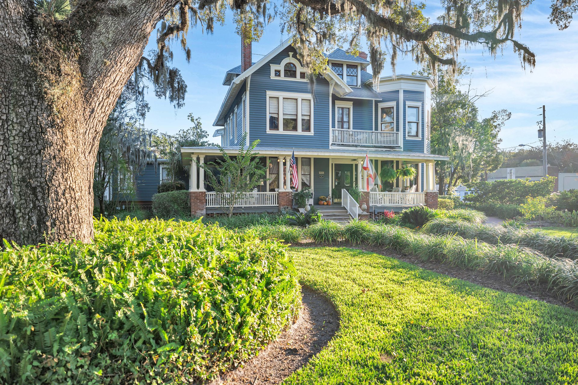 A large blue Victorian-style house with a wraparound porch sits behind lush green landscaping and a curved walkway. Mature trees with hanging moss frame the scene on a sunny day.