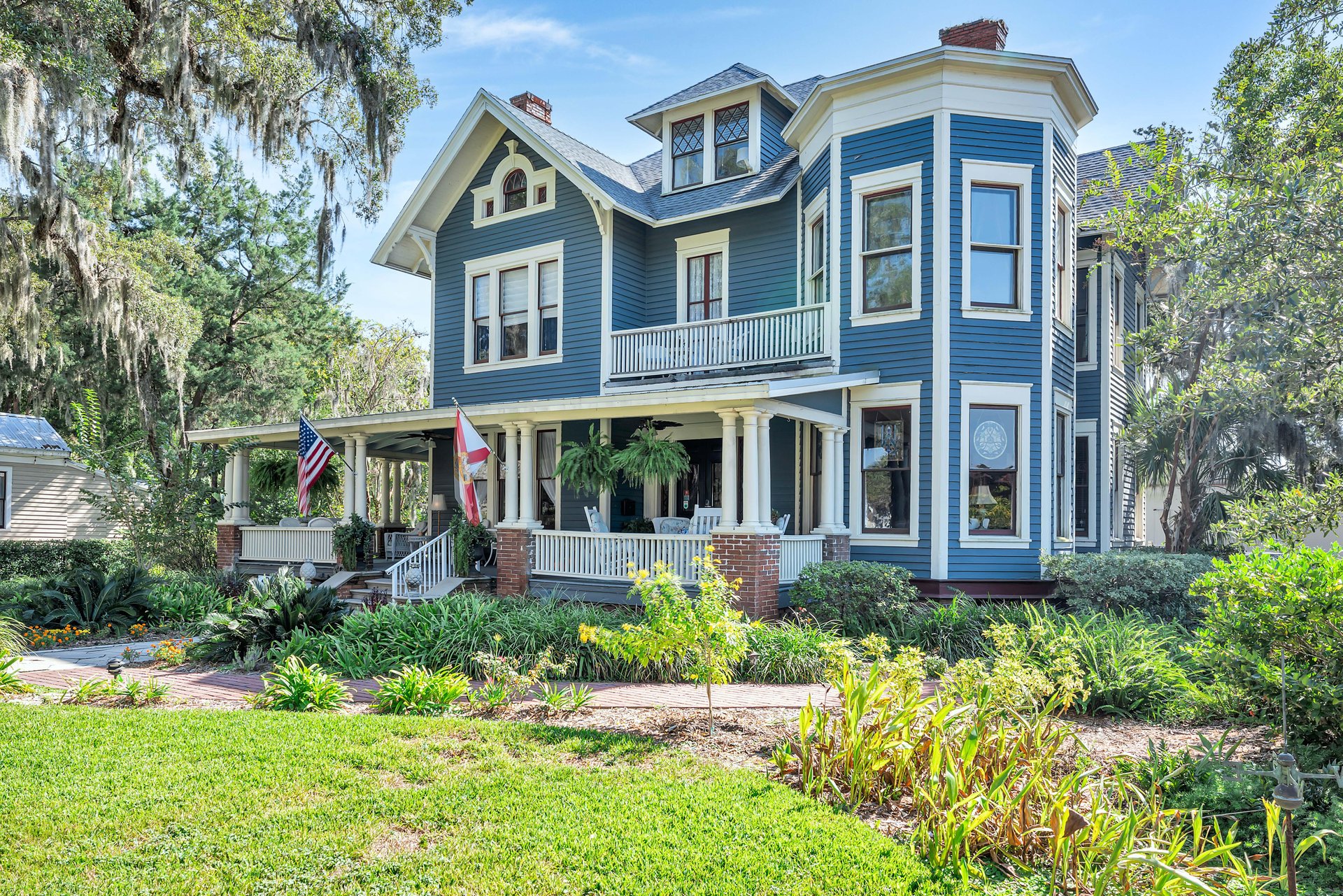 A large, blue Victorian-style house with white trim, a wraparound porch, American and state flags, and lush landscaping, surrounded by trees under a clear sky.