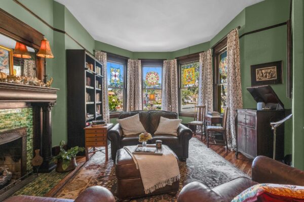A cozy living room with green walls, stained glass bay windows, a dark wood fireplace, bookshelves, armchairs, an ottoman, and vintage-style furnishings. Natural light fills the space, creating a warm atmosphere.