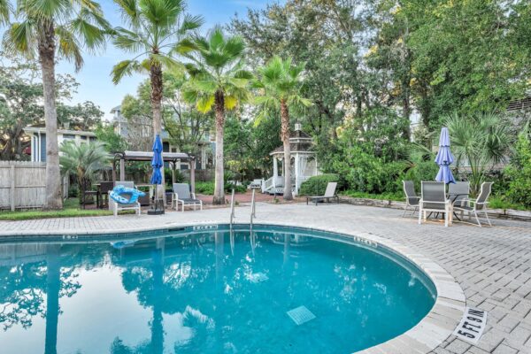 Curved outdoor swimming pool surrounded by palm trees, lounge chairs, umbrellas, a gazebo, and a patio table with chairs, set in a lush, green backyard with privacy fencing.