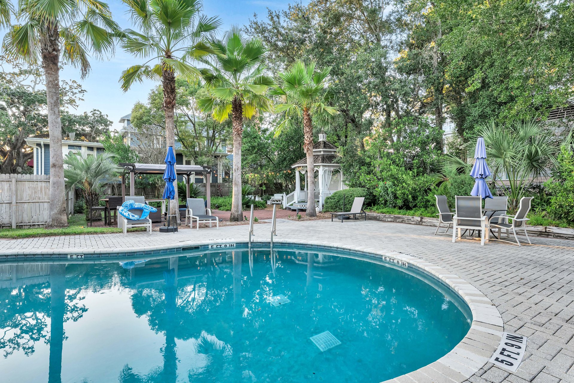 Curved outdoor swimming pool surrounded by palm trees, lounge chairs, umbrellas, a gazebo, and a patio table with chairs, set in a lush, green backyard with privacy fencing.