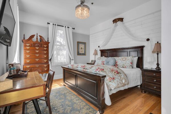 Elegant bedroom with a dark wood bed, floral bedding, two lamps, a wooden chest of drawers, a desk with a chair, a blue rug, white curtains, and a decorative canopy above the bed.