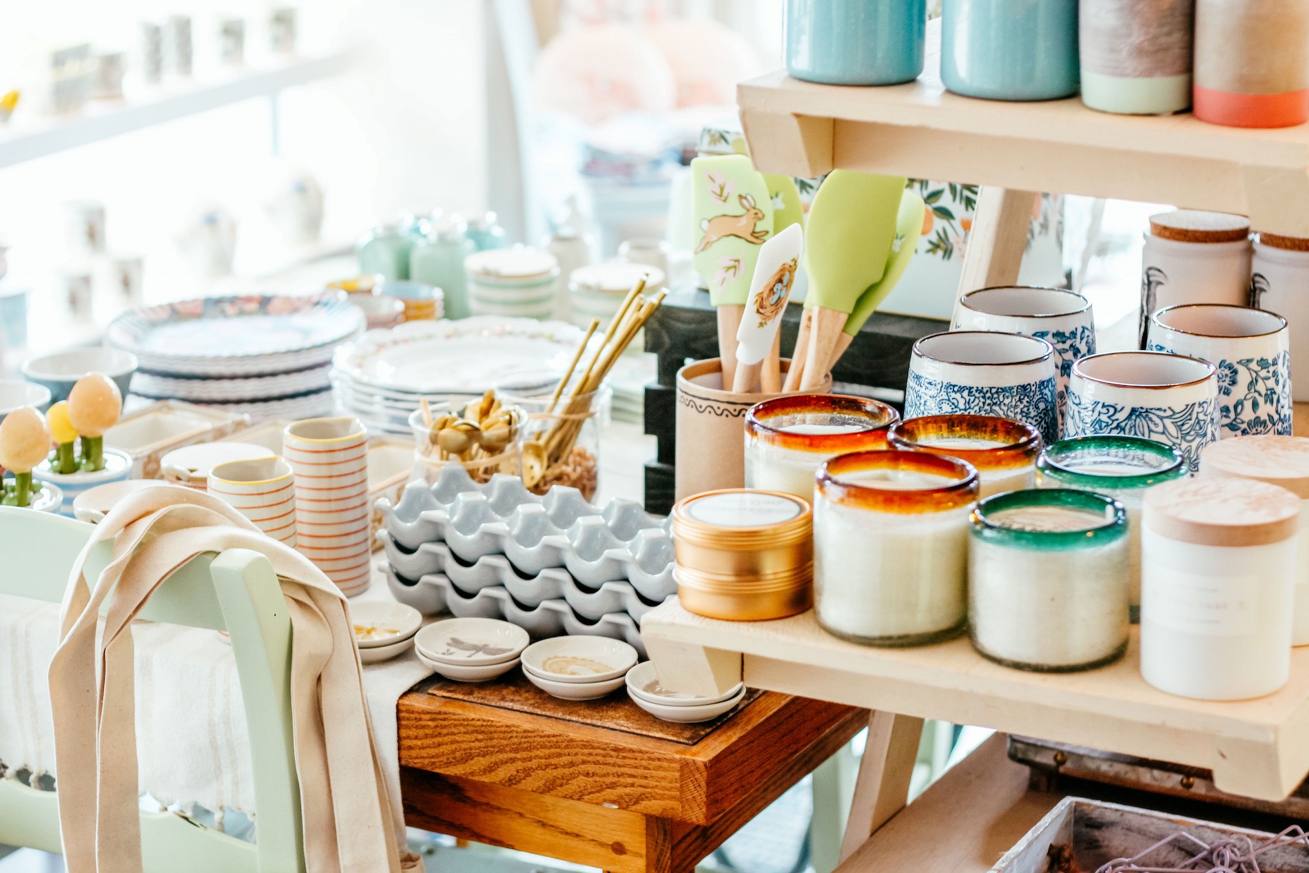 A bright display of assorted home goods including stacked plates, cups, candles, utensils, egg trays, and kitchen accessories arranged neatly on wooden shelves and tables in a well-lit shop.