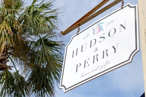 A white hanging sign with Hudson & Perry home and gifts and est 2016 in black text, featuring a peach illustration. A palm tree and blue sky are visible in the background.
