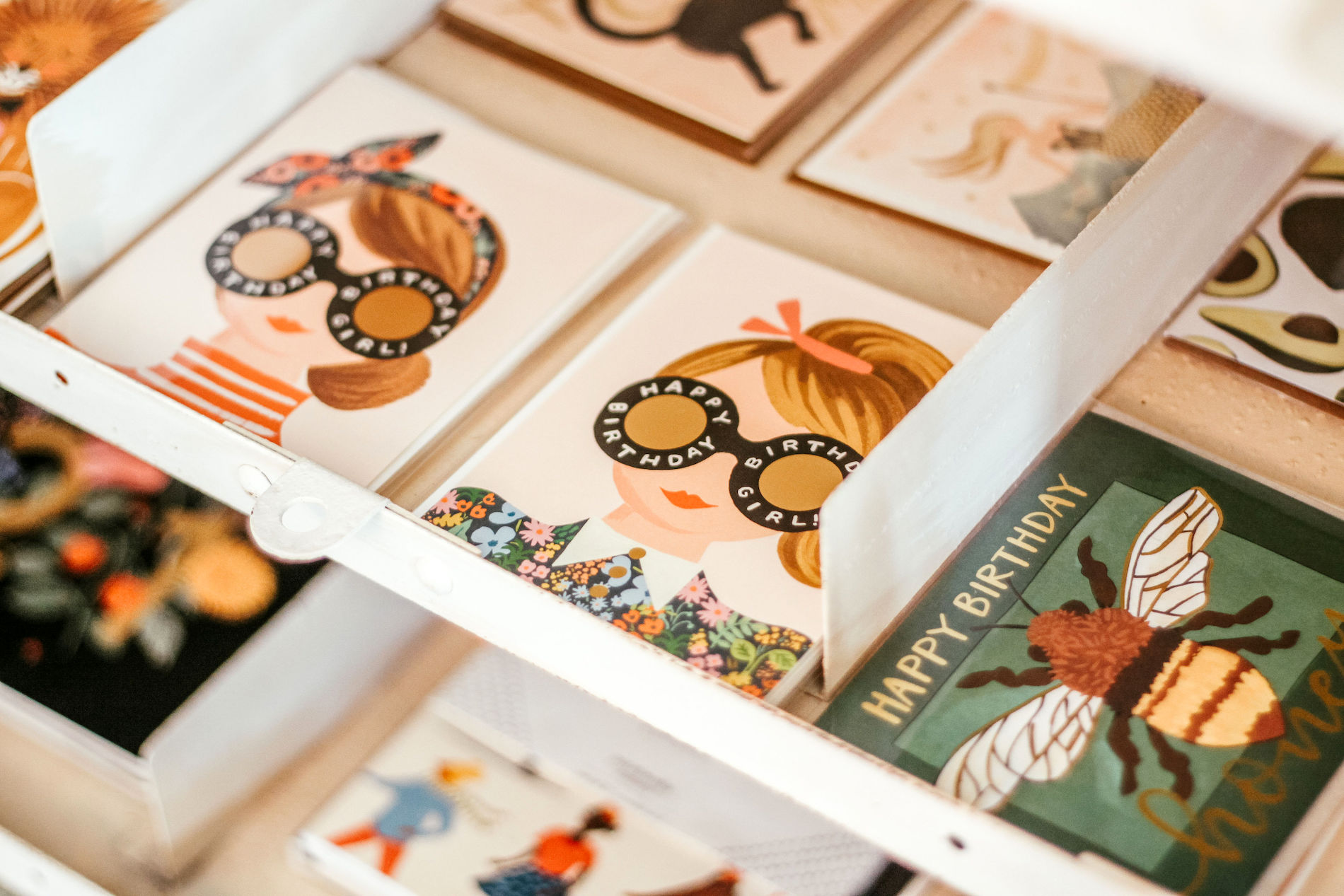 A display shelf with various illustrated greeting cards, including birthday cards featuring women with Happy Birthday Girl glasses and a card with a bee illustration.