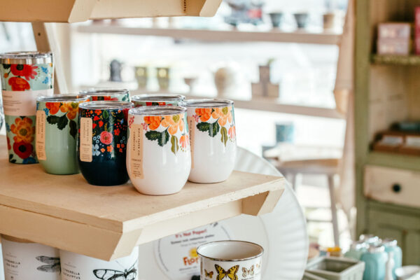 Several colorful, floral-patterned insulated tumblers are displayed on a wooden shelf in a bright, sunlit shop with various items in the background.