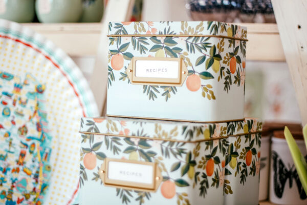 Two decorative metal boxes labeled RECIPES are stacked on a shelf. The boxes have a floral and fruit pattern with green leaves and orange fruits. Other kitchen items are visible in the background.