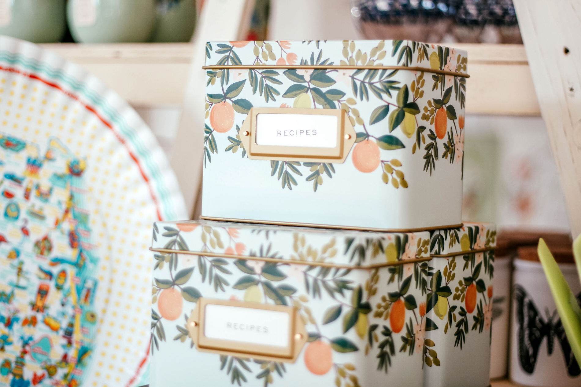 Two decorative metal boxes labeled RECIPES are stacked on a shelf. The boxes have a floral and fruit pattern with green leaves and orange fruits. Other kitchen items are visible in the background.