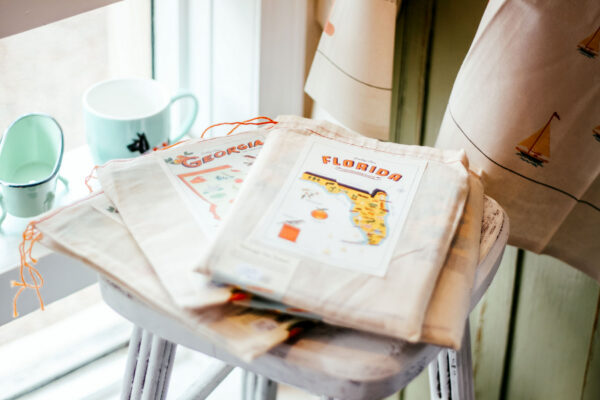 Cloth bags with vintage state-themed prints, including Florida and Georgia, are displayed on a white stool near a window. Two light green cups and a curtain with sailboat designs are in the background.