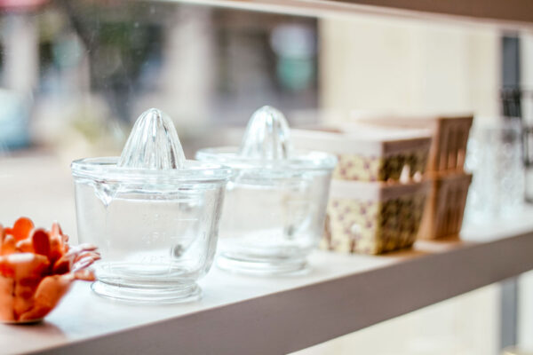 Two clear glass citrus juicers sit on a white windowsill next to small decorative containers and orange ceramic objects, with soft daylight streaming through the window.