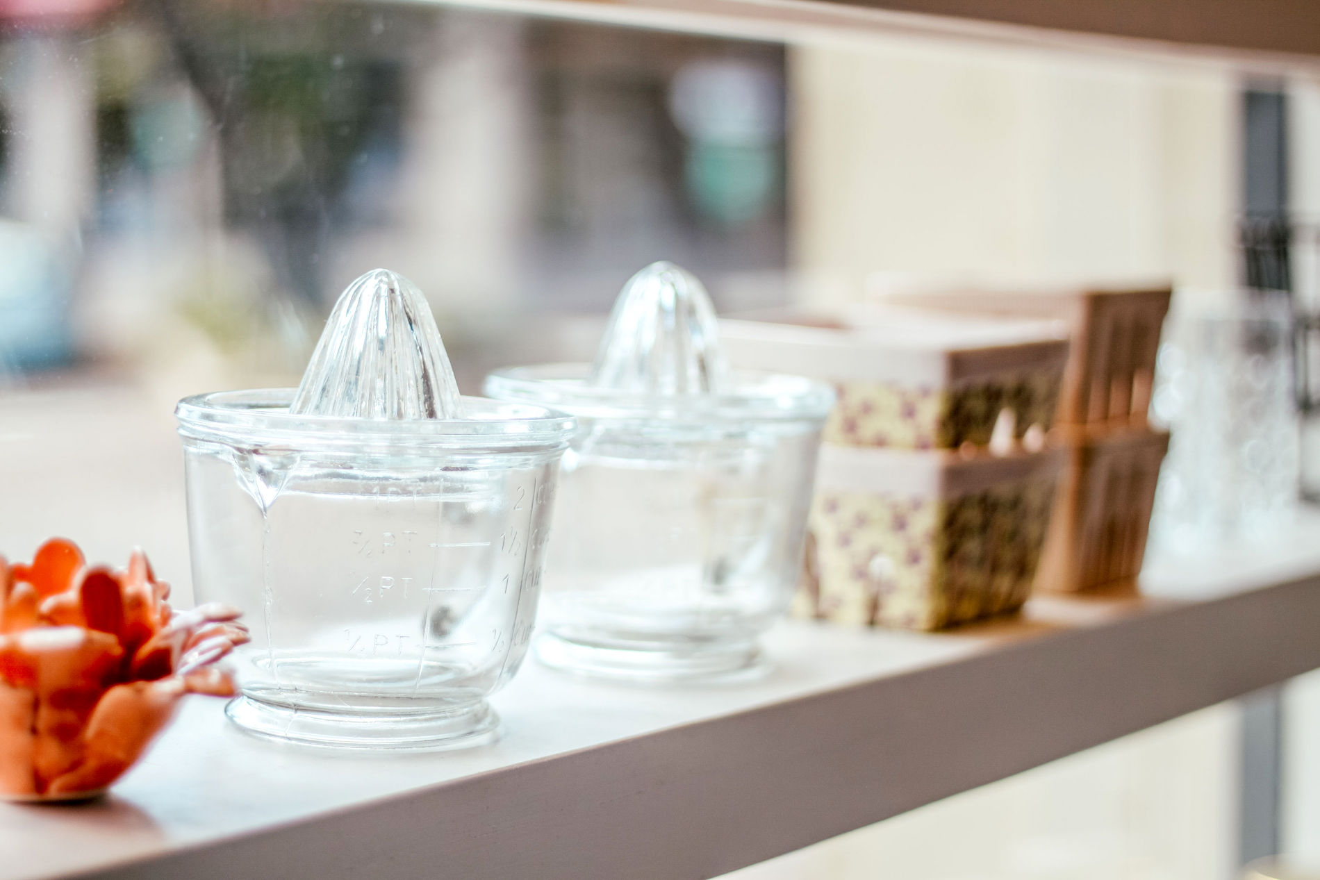 Two clear glass citrus juicers sit on a white windowsill next to small decorative containers and orange ceramic objects, with soft daylight streaming through the window.