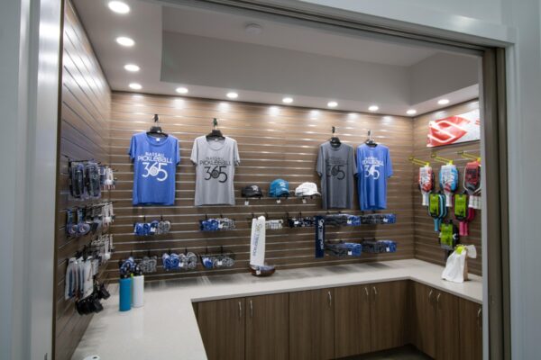 A retail display with blue and gray pickleball shirts, hats, water bottles, and colorful pickleball paddles hanging on a wall above a light countertop with cabinets below.