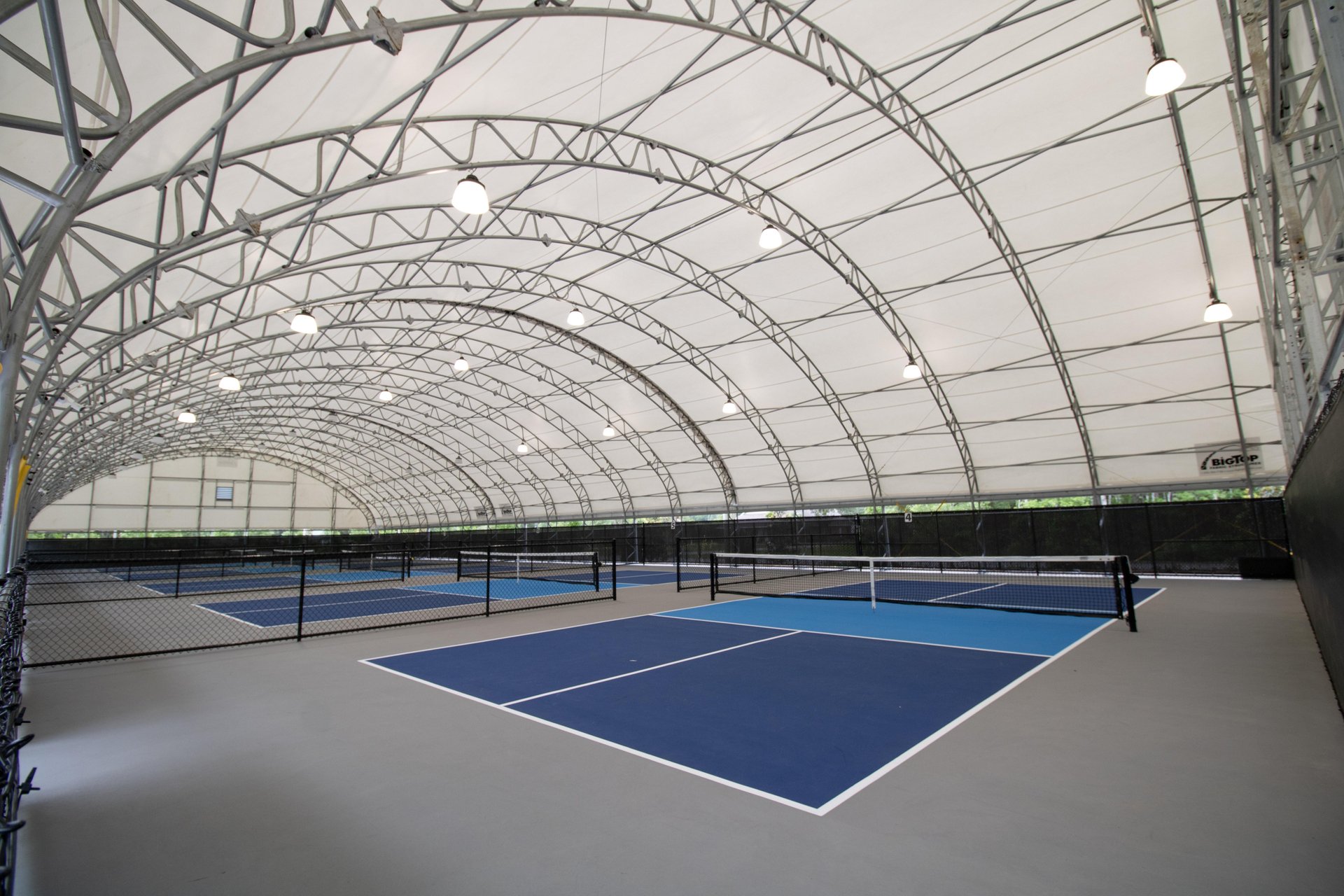 An indoor sports facility with arched metal beams and a white canopy roof, featuring multiple blue and gray tennis or pickleball courts separated by black fences. Bright overhead lights illuminate the space.