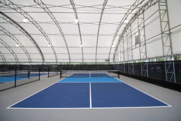 Indoor tennis courts with blue playing surfaces and white boundary lines, separated by black nets under a large white canopy with overhead lights and metal framework. The courts are empty.