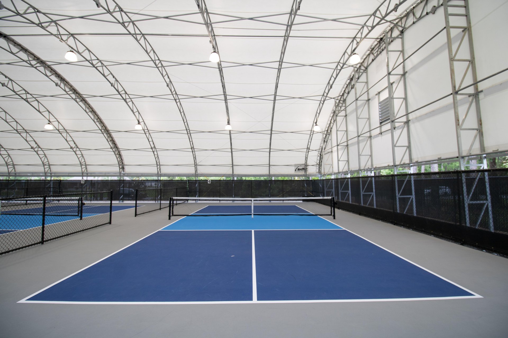 Indoor tennis courts with blue playing surfaces and white boundary lines, separated by black nets under a large white canopy with overhead lights and metal framework. The courts are empty.