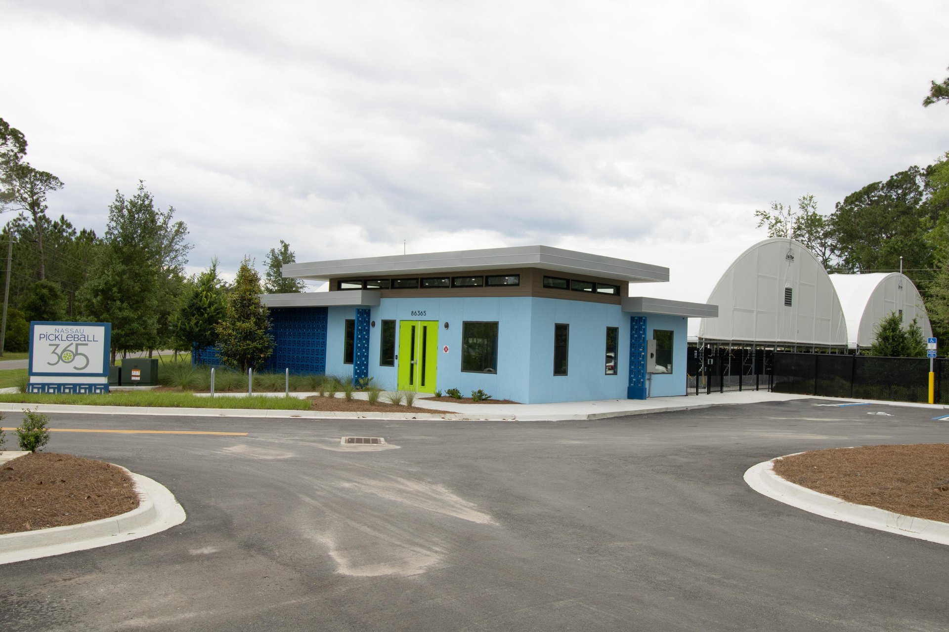 A small, modern light-blue building with a flat roof and green door sits in front of two large white domes. A sign reads Pickleball 365. The area is surrounded by trees and a newly paved driveway.