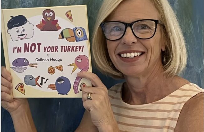 A smiling woman with blonde hair and glasses holds up a childrens book titled IM NOT YOUR TURKEY! by Colleen Hodge. The book cover features illustrated turkeys and food items.